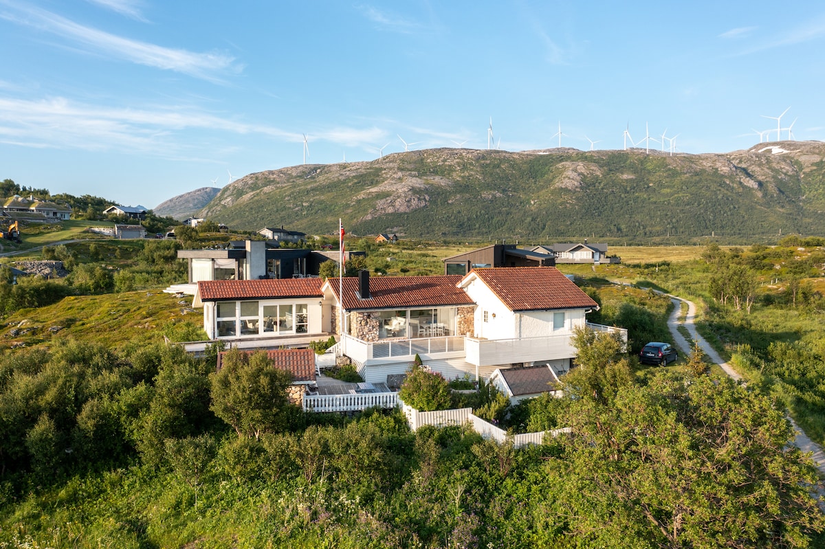 A spacious house is set against a backdrop of rolling hills and wind turbines, surrounded by lush greenery. The architecture features a mix of traditional and modern elements, with a tile roof and white-washed walls. A driveway leads to the property, providing easy access.
