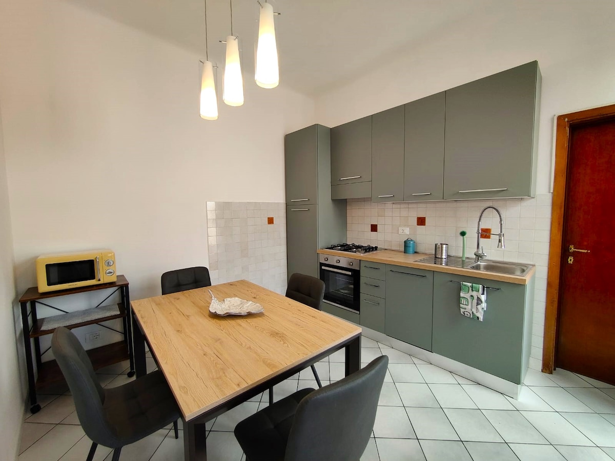 A modern kitchen area is highlighted by a light wooden table surrounded by black chairs. Built-in cabinets and appliances, including a microwave and gas stove, provide a functional cooking space. Bright pendant lights hang above, contributing to a well-lit atmosphere.