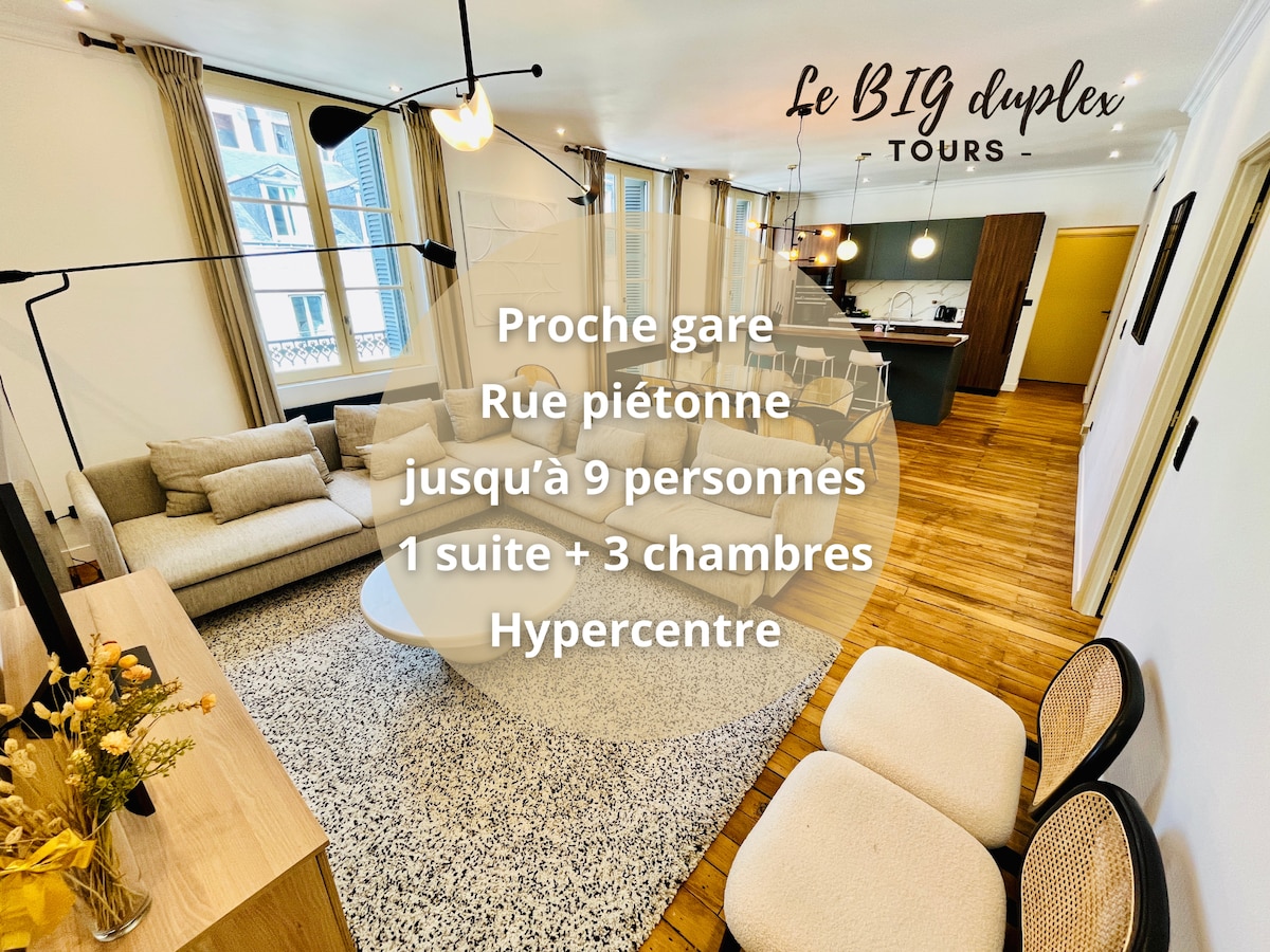 The living area features a spacious layout with a comfortable sofa and two armchairs. A coffee table and a low buffet with a flat-screen TV are positioned within the bright space, complemented by warm wooden flooring and large windows inviting natural light.