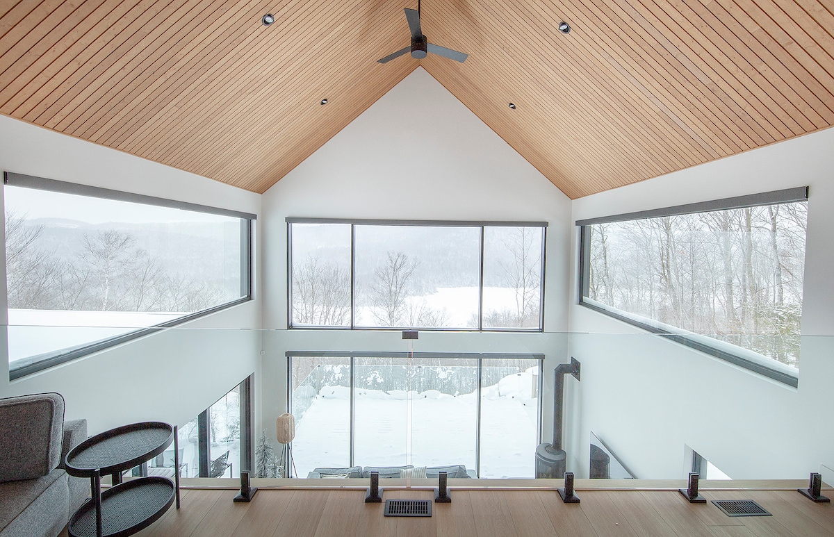 A spacious living area features large windows that frame views of a snowy landscape, allowing natural light to fill the space. A wooden ceiling adds warmth to the contemporary design, while modern furnishings provide comfort in the open layout.