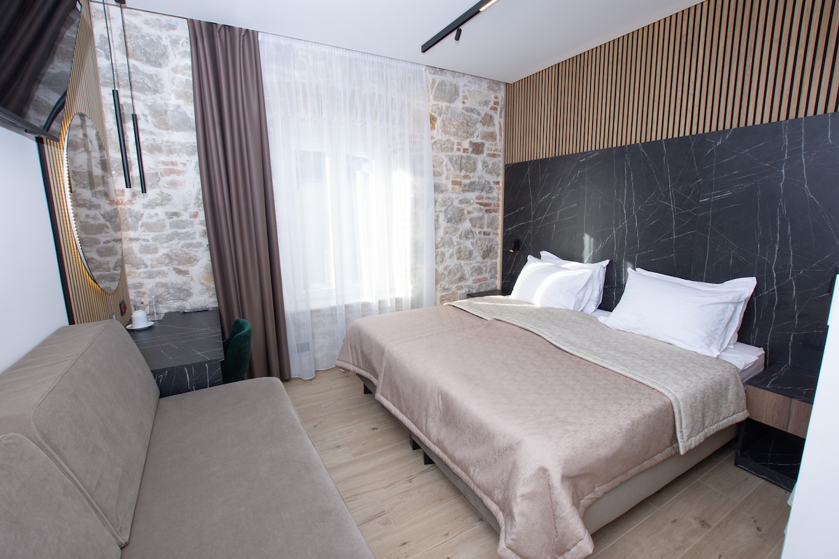 A modern deluxe room features a spacious bed with a light-colored quilt draped over it. A comfortable seating area is visible beside the bed. Natural light filters through sheer curtains, illuminating the stone and wood accents on the walls.
