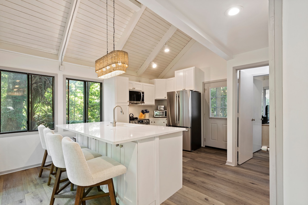 The kitchen area features a spacious island with three high-backed chairs and a modern light fixture overhead. Large windows allow natural light to fill the space, highlighting the sleek countertops and stainless steel appliances. The open layout connects to adjoining rooms, providing a bright and airy ambiance.