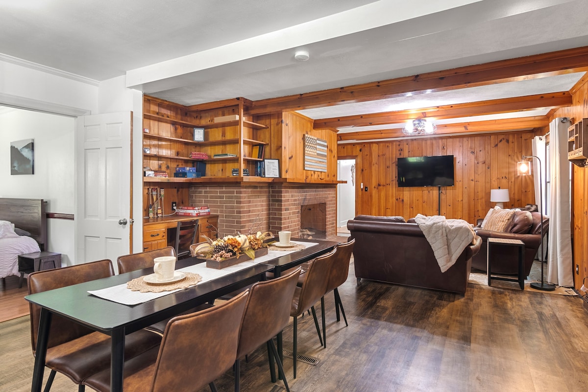 An open living and dining area features a long dining table set with decorative elements. A comfortable sofa sits nearby, facing a mounted television. Warm wooden paneling adds a natural touch, while shelves and a fireplace contribute to the cozy ambiance.