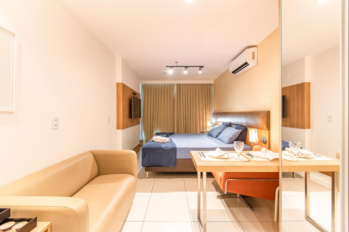 A spacious studio layout is presented with a comfortable sofa and dining area. A large bed is visible in the background, with soft linens and pillows. The setting is illuminated by natural light flowing through floor-to-ceiling curtains, and a television is mounted on the wall.