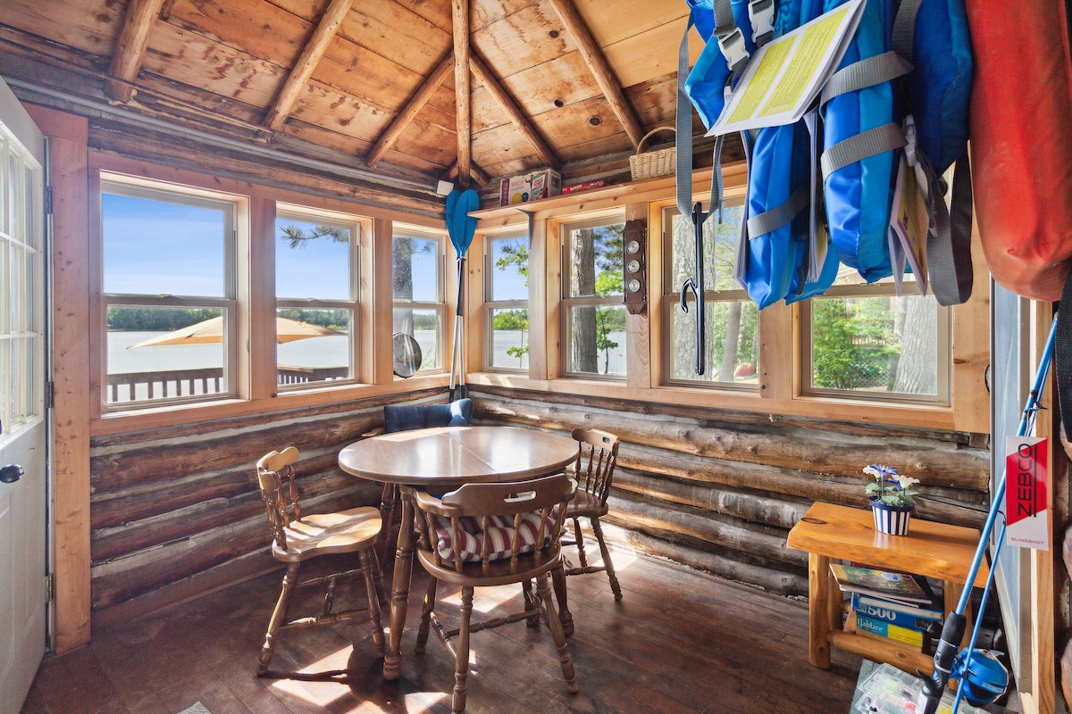 A bright dining area features a round wooden table surrounded by four chairs, complemented by large windows allowing natural light to brighten the space. The walls are constructed of logs, and decorative items are neatly arranged on nearby shelves. Outdoor views of Snyder Lake are visible through the windows.