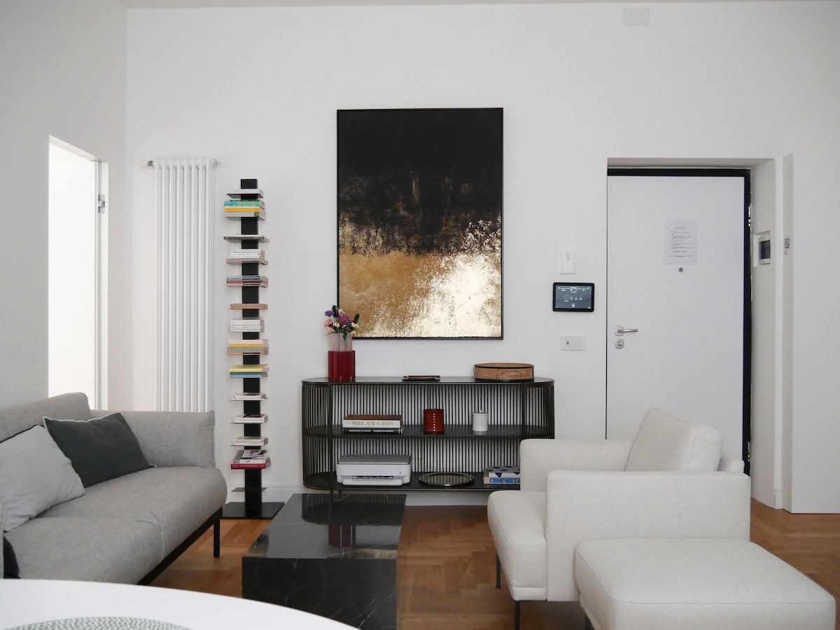 A modern living room features a neutral color palette with plush sofas and a sleek coffee table. A large abstract artwork dominates the wall, complemented by a bookshelf filled with books. The space is well-lit, enhancing the minimalist design.