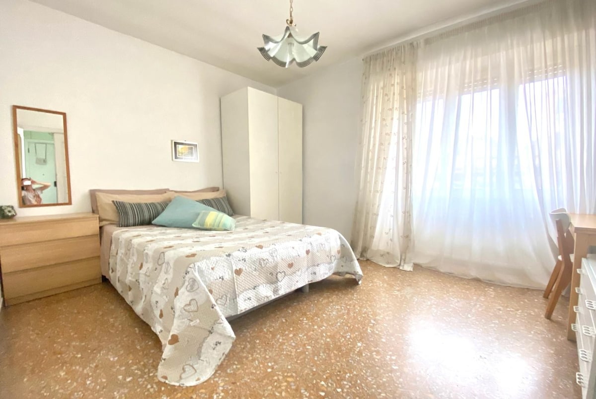 A well-lit bedroom features a spacious bed with a patterned bedspread and decorative cushions. Natural light streams through a large window, which is framed by light, airy curtains. A simple wooden wardrobe and a small desk are positioned alongside the bed, contributing to the room's functionality.