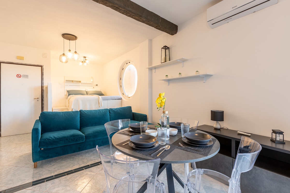 A modern open space features a cozy seating area with a teal sofa, accompanied by a round dining table set for four. The elegantly designed lofted bed is visible in the background, complemented by circular windows. Soft lighting enhances the welcoming ambiance.