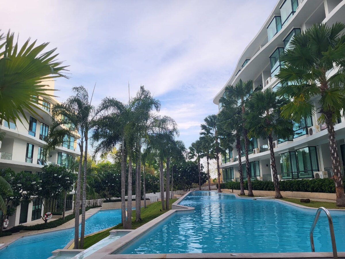 A serene swimming pool is surrounded by modern buildings, featuring palm trees that provide a tropical atmosphere. The clear water reflects the blue sky, while pathways and landscaped greenery enhance the tranquil setting.