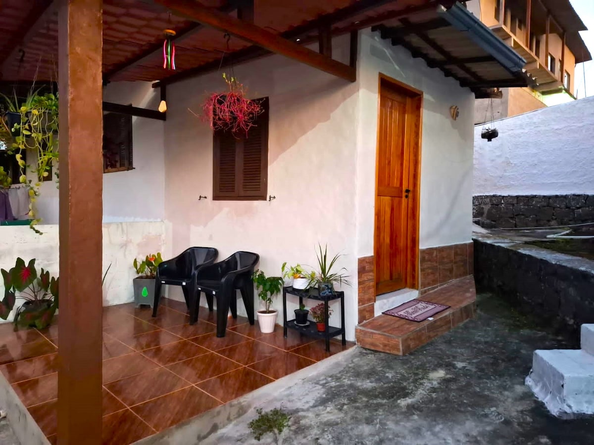 An outdoor seating area is visible, featuring two black chairs positioned on a tiled patio. A small potted plant sits on a stand beside the chairs. The entrance to the accommodation is marked by a wooden door with a decorative welcome mat, while plants and hanging decorations add a touch of greenery.