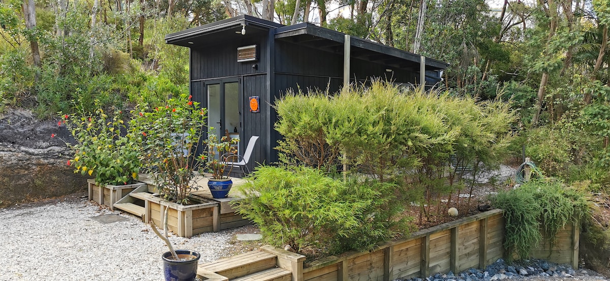 A modern cabin is nestled within a lush natural landscape, surrounded by a variety of green shrubs and colorful flowers. Gravel pathways lead to the entrance, where large windows provide an inviting glimpse of the interior.