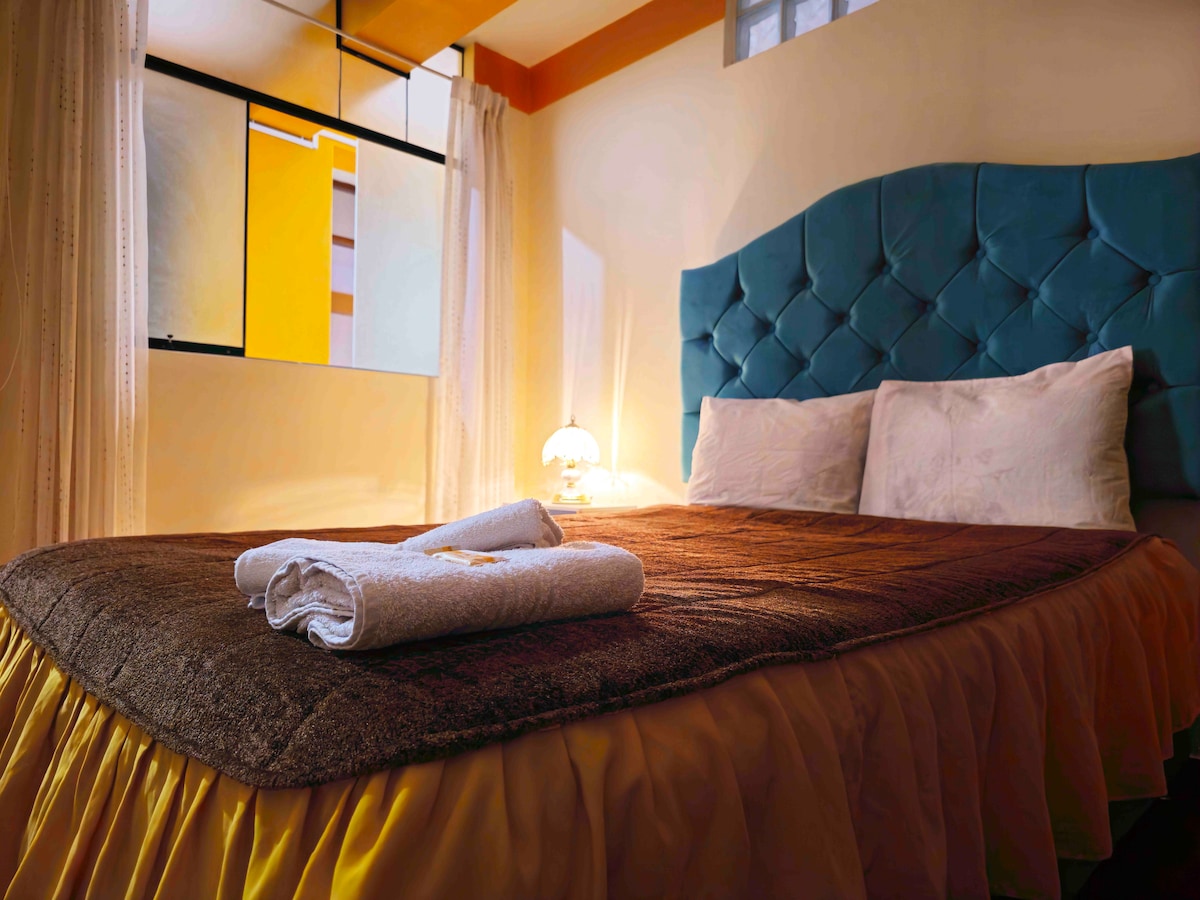 A spacious bed with a tufted turquoise headboard is featured, dressed in neutral-toned linens. Two neatly rolled towels rest on the bedspread. Light filters through sheer curtains, highlighting the warm colors of the room.