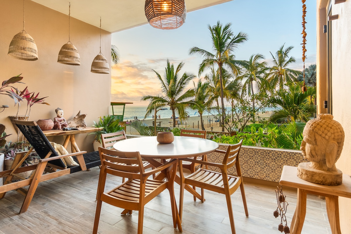 An outdoor balcony area features a round dining table surrounded by wooden chairs. Pendant lights hang from the ceiling, while tropical plants and a vibrant view of palm trees and the ocean are visible in the background, creating a serene setting.