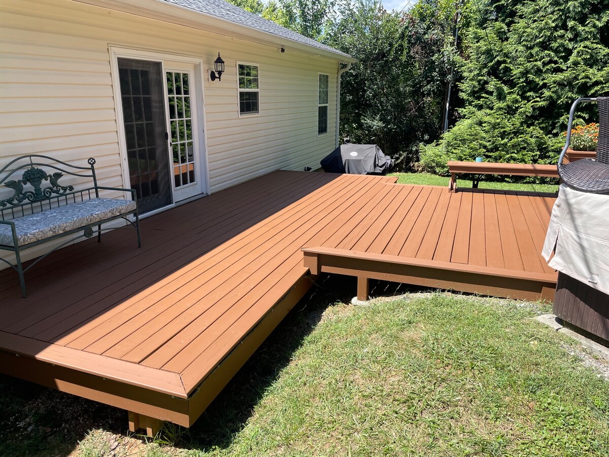 A spacious wooden deck offers ample room for relaxation and outdoor activities. A comfortable bench is positioned beside the house, while a hot tub is visible in the corner. Lush greenery surrounds the area, providing a peaceful atmosphere.