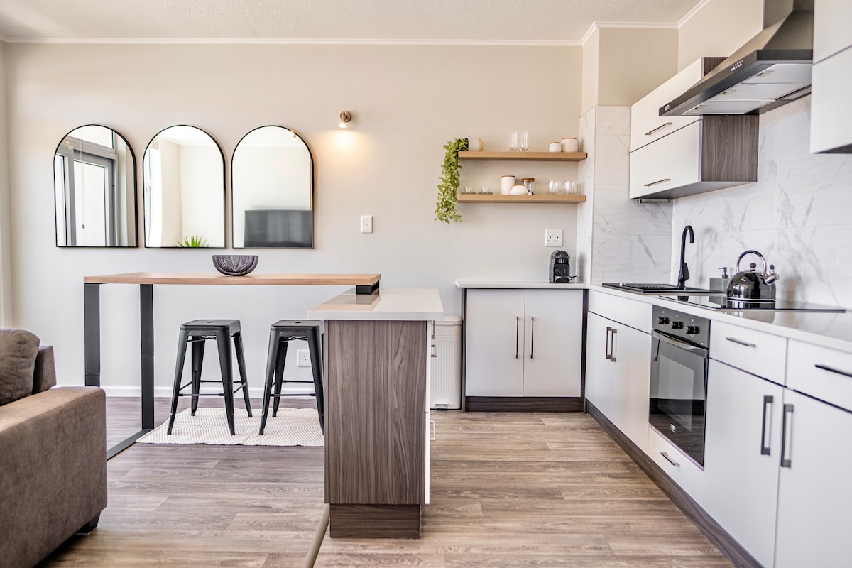 The modern kitchen and dining area create a seamless space. A sleek kitchen island is paired with two black stools, while the kitchenette features contemporary appliances and minimalist cabinetry. Three round mirrors and open shelving enhance the airy, inviting atmosphere.