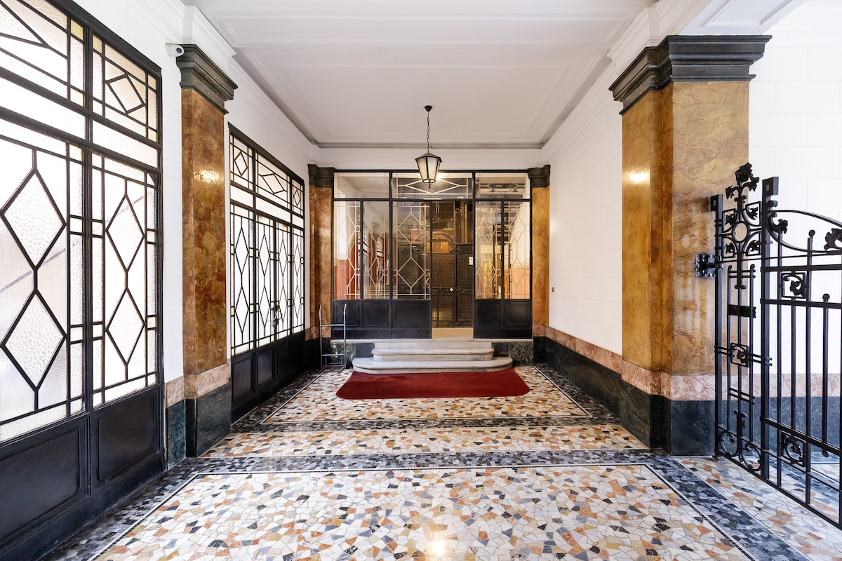 An elegant entrance hall features ornate marble flooring with geometric patterns. Tall windows allow natural light to fill the space, illuminating rich marble columns and decorative details. A grand staircase is positioned at the back, leading to the elevator access and lobby area.