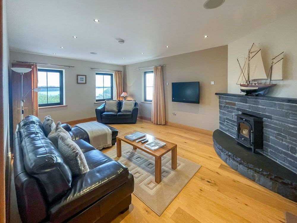 The living room features two comfortable leather sofas and a wooden coffee table. A flat-screen TV is mounted on the wall, while a faux stove adds a cozy touch. Large windows provide views overlooking Donegal Bay, with soft curtains framing the natural light.