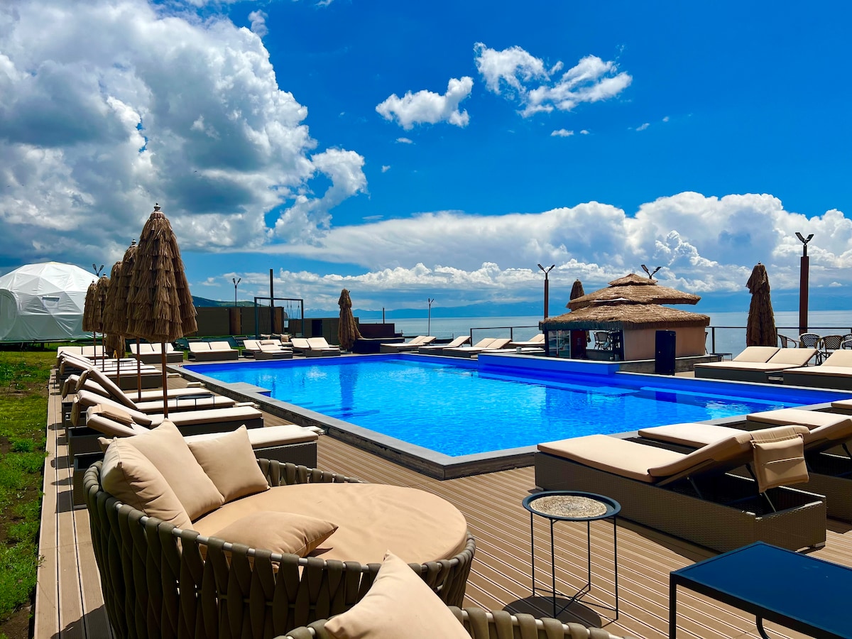An outdoor pool is surrounded by sun loungers and shaded seating areas. Palms and umbrellas provide comfort, while a thatched-roof bar offers refreshments. The striking blue water of the pool contrasts with the lush green landscape and is framed by a bright sky with scattered clouds.