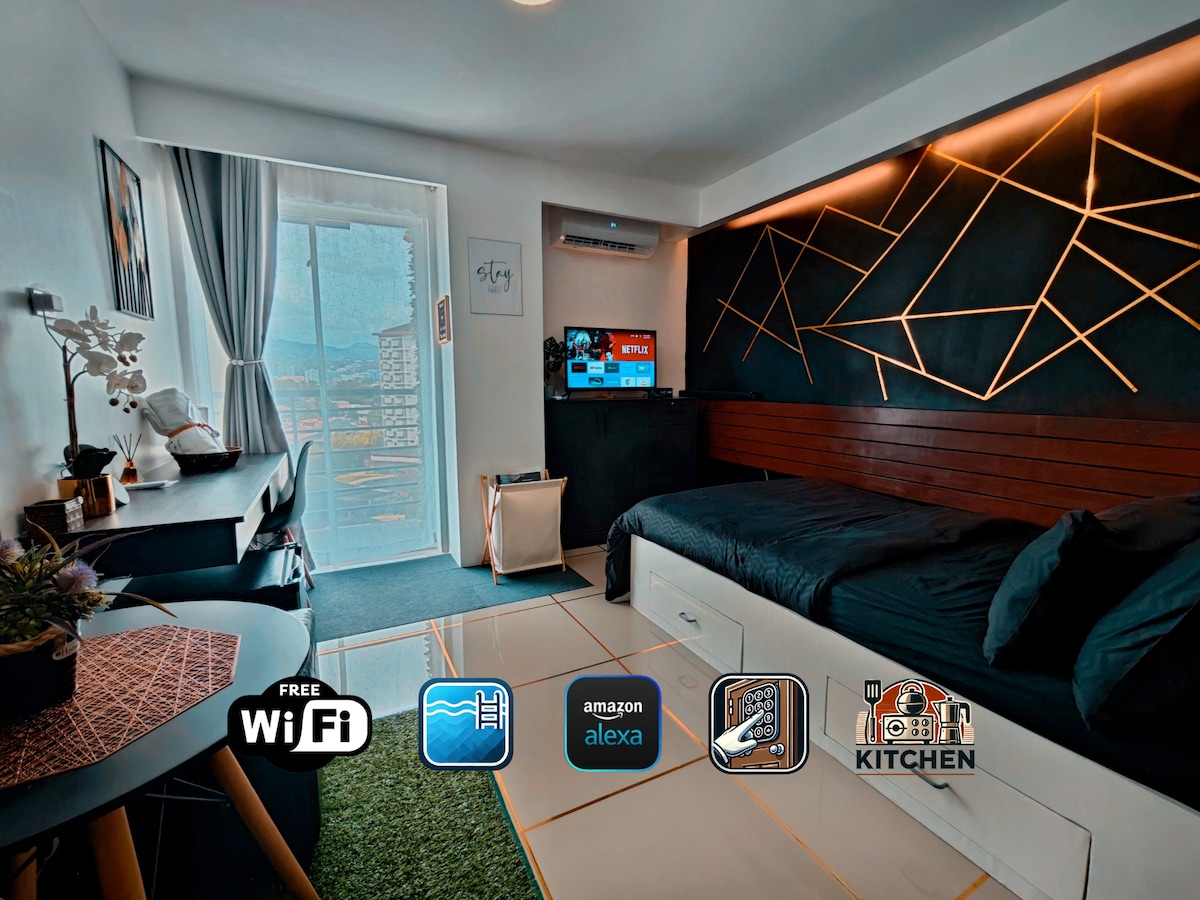 A modern studio features a spacious bed against a stylish black and wooden backdrop. A smart TV is mounted on the wall opposite the bed, while a compact work station sits beside a large window, which allows natural light to illuminate the space. Wi-Fi and kitchen symbols are depicted.