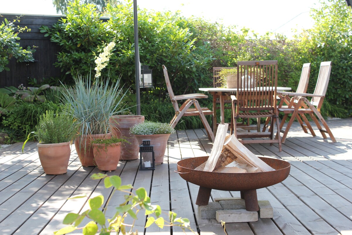 An outdoor terrace is surrounded by greenery, featuring a table and six wooden chairs. Various potted plants are arranged throughout the area, and a fire pit with stacked wood is positioned centrally, adding to the relaxing outdoor ambiance.