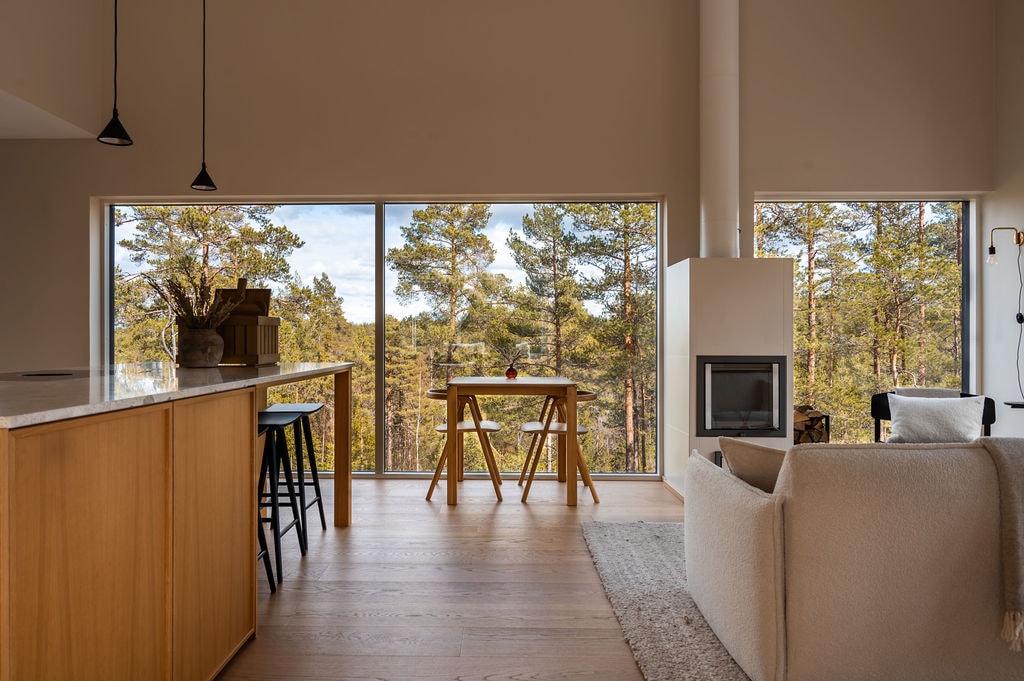An open-plan living area features a large window overlooking the surrounding forest. A cozy sofa is positioned near a modern fireplace, while a simple dining table for two is set beside the kitchen island. Neutral tones enhance the calming atmosphere.