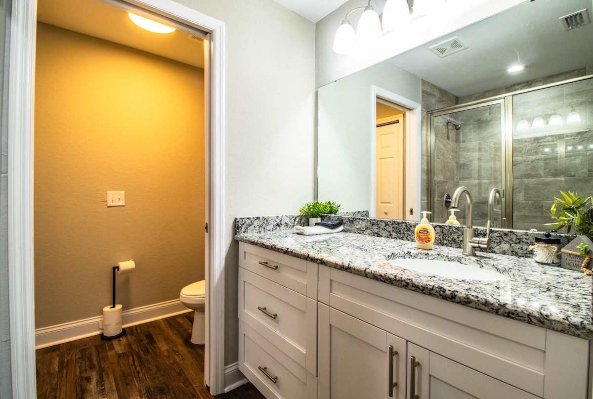 A modern bathroom features a dual sink countertop with a granite surface. A spacious shower enclosure is visible, fitted with glass doors. A neutral-toned wall complements the warm lighting, and a partially open door leads to a separate toilet area.