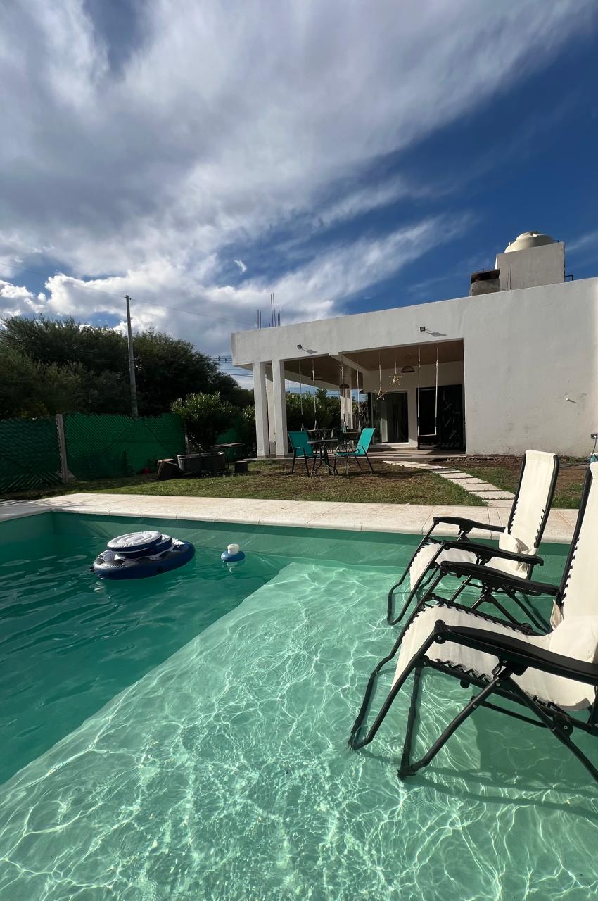 A serene outdoor area is shown, featuring a clear swimming pool with lounge chairs positioned nearby. The backdrop includes a spacious white structure with large windows, surrounded by greenery. The sky is partly cloudy, lending a calm atmosphere to the scene.