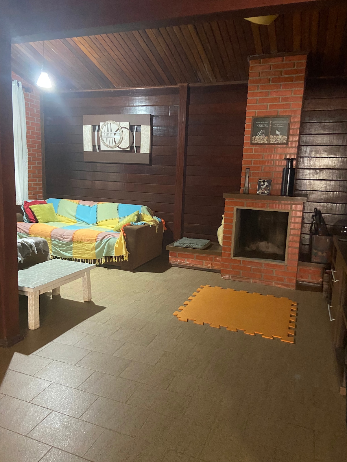 An inviting living area is outlined by wooden paneling and a cozy sofa adorned with a colorful throw. A brick fireplace is visible, providing a warm focal point, while a small seating area features a simple bench. Light enters through a nearby window, enhancing the space.