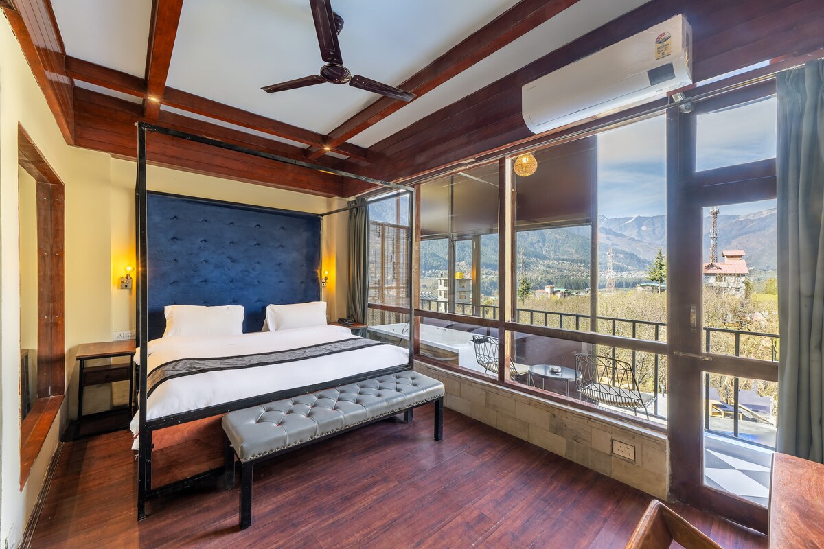 A spacious room features a large bed with a plush headboard and a chic bench at the foot. Floor-to-ceiling windows invite natural light and offer stunning mountain views. Elegant wooden beams and warm wood tones contribute to the inviting ambiance.