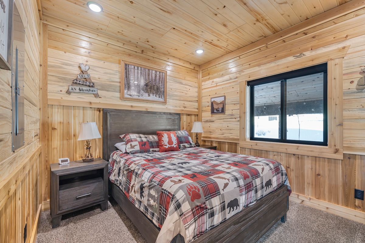 Een gezellige slaapkamer met een queensize bed met zacht beddengoed en een warme inrichting in hutstijl. De kamer is voorzien van een aan de muur gemonteerde tv, dressoir en met hout beklede muren die een comfortabele en uitnodigende bergretraite creëren. 