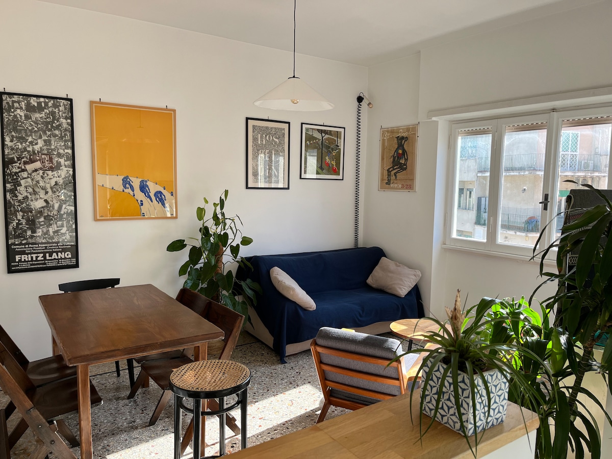 A bright dining area includes a wooden table surrounded by chairs, with a sofa positioned nearby. Artwork adorns the walls, complemented by indoor plants. Natural light enters through windows, creating a warm atmosphere in the space.