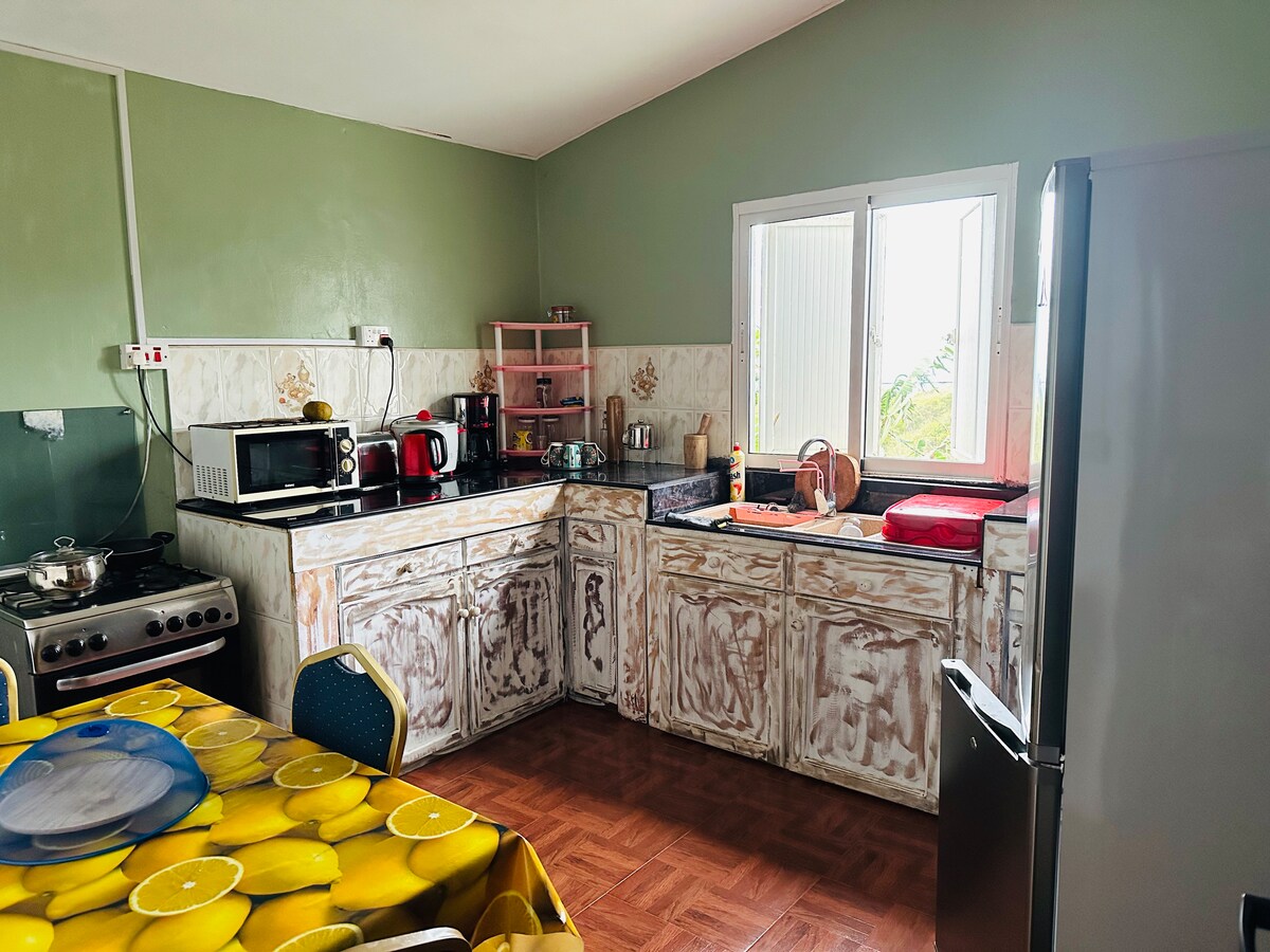 The spacious kitchen features a combination of essential appliances, including a microwave, stove, and refrigerator. Natural light fills the area through a window above the sink. A dining table adorned with a lemon-patterned cloth adds a pop of color to the neutral walls and cabinets.