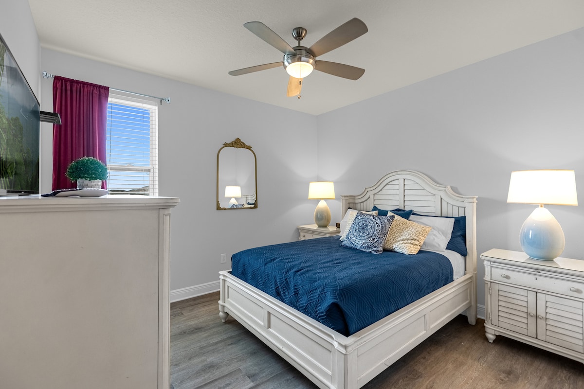 חדר שינה אלגנטי עם מיטה נוחה מוקפת בשני אהילים מסוגננים על מגירות תואמות. נברשת אופנתית תלויה מעל, ומשקפת מראה על הקיר. טלוויזיה עם מסך שטוח יושבת מעל מגירה אלגנטית, עם חלון מואר שמשלים את החלל השלו.