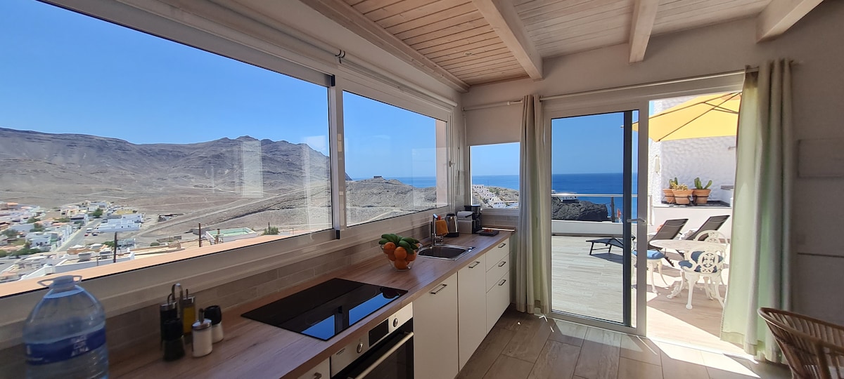 A bright kitchen is visible, featuring a wooden countertop and modern cooking appliances. Large windows allow for natural light and offer stunning views of the ocean and hills. An open door leads to a terrace, where outdoor seating and an umbrella are seen.