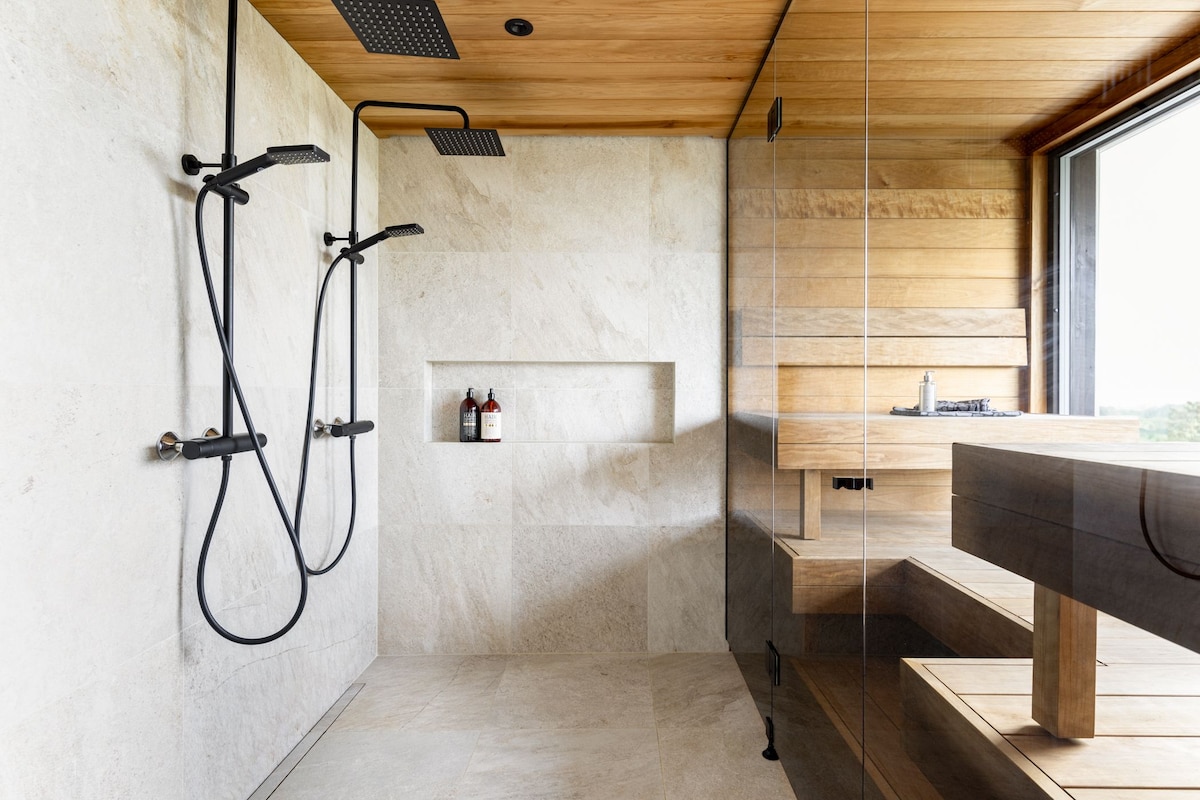 A modern bathroom features a spacious glass shower area equipped with two black showerheads. Light-colored stone walls complement natural wood accents. A wooden sauna bench and steps add warmth, while a shelf displays bath products. A large window offers scenic views and natural light.