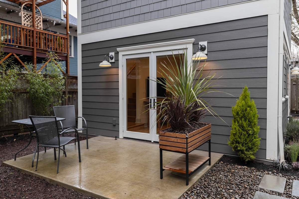 A private patio area features a smooth concrete surface surrounded by landscaping, including a potted plant and small shrub. Two patio chairs and a glass table are arranged for outdoor seating, while double french doors lead into the interior of the guesthouse.