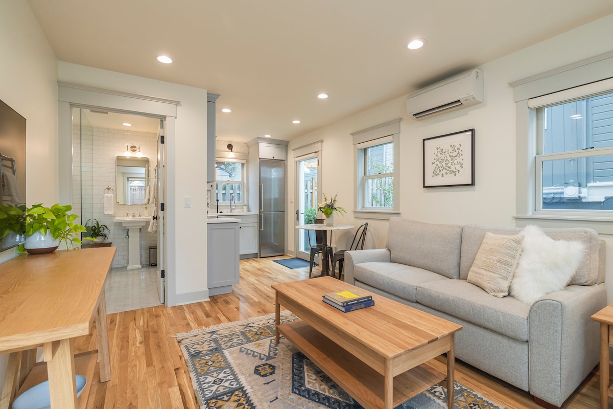 A light-filled living area showcases a comfortable gray sofa and a wooden coffee table, complemented by a decorative rug. The open layout reveals a dining table set for two. A hallway leads to modern bathrooms with accessible features, enhancing the welcoming atmosphere.