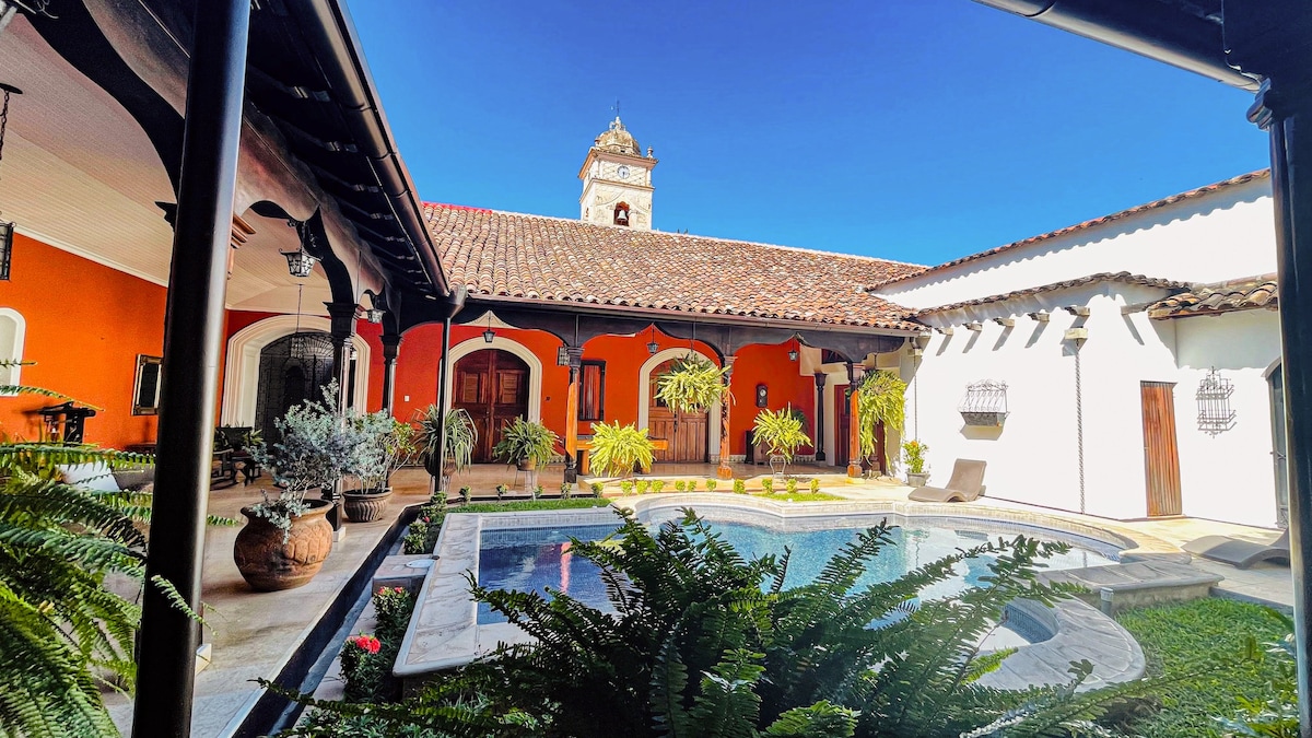 An inviting courtyard is surrounded by adobe walls and elegant arches, with a serene pool at its center. Lush green plants and decorative pottery enhance the tranquil atmosphere, while the backdrop features a colonial tower, offering a glimpse of the sky above.