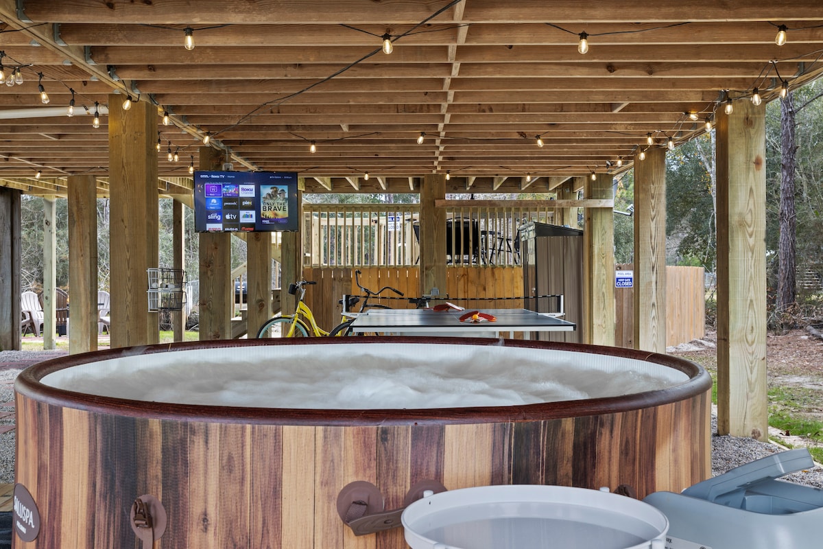 A wooden hot tub is positioned under a covered area, surrounded by string lights. A flat-screen TV is mounted on a nearby wall. Bicycles and a ping pong table are visible in the background, providing options for outdoor entertainment.