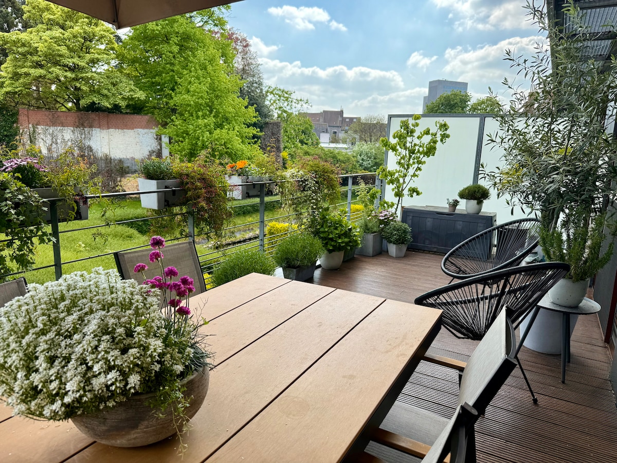 A spacious terrace is presented, featuring a wooden dining table surrounded by comfortable seating. Potted plants add greenery, while a backdrop of lush vegetation and trees creates a serene outdoor space, complemented by a clear blue sky dotted with clouds.