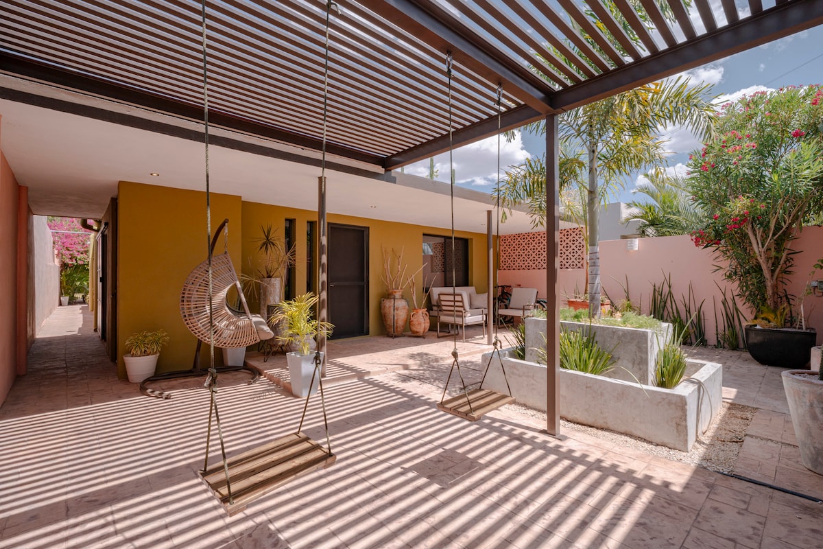 An outdoor area features two hanging swings suspended from a structured overhead, providing a comfortable space for relaxation. Planters with greenery frame the space, along with a serene garden area featuring low stone planters. Natural light filters through, creating a calm atmosphere.