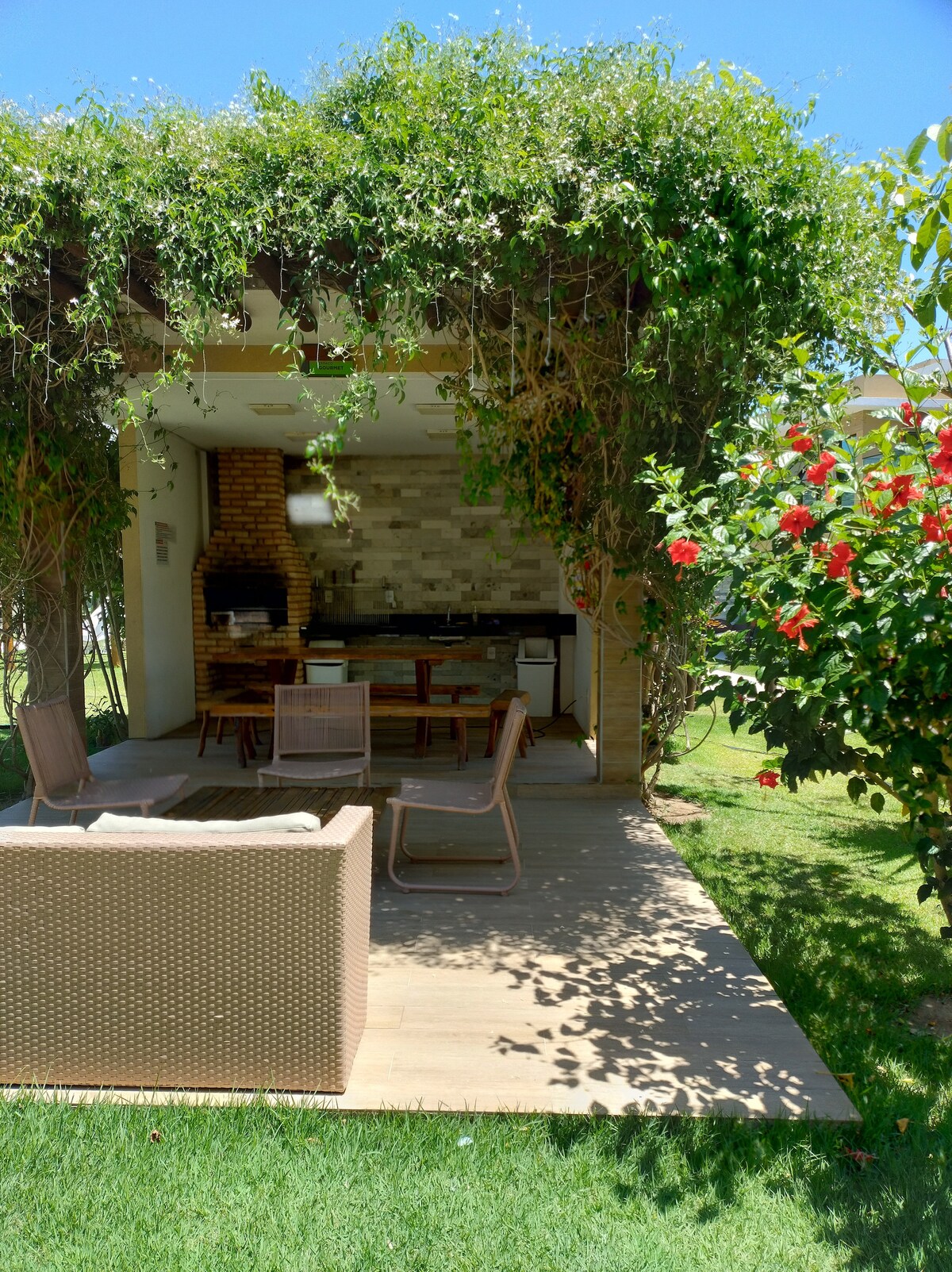 An outdoor space is shown, featuring a covered dining area with a rustic brick barbecue. Surrounding greenery enhances the inviting atmosphere, with a blend of flowering plants visible. Comfortable seating is arranged on a wooden deck, providing a relaxed setting for gatherings.