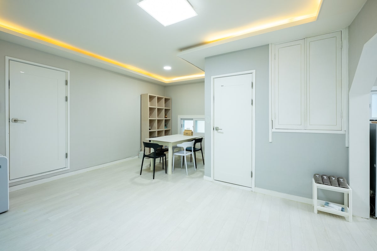 A bright, spacious room features light-colored flooring and walls, complemented by soft ambient lighting. A simple dining table with four chairs occupies the center, while a shelving unit stands against one wall. Two doors are visible, adding functional access to the space.