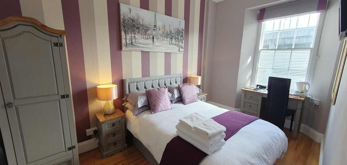 A well-appointed bedroom features a spacious bed with neatly arranged linens and towels. Soft purple accents adorn the walls and bedding, while bedside lamps provide warm lighting. A small desk with a chair is situated nearby, complemented by a tasteful piece of artwork above the bed.