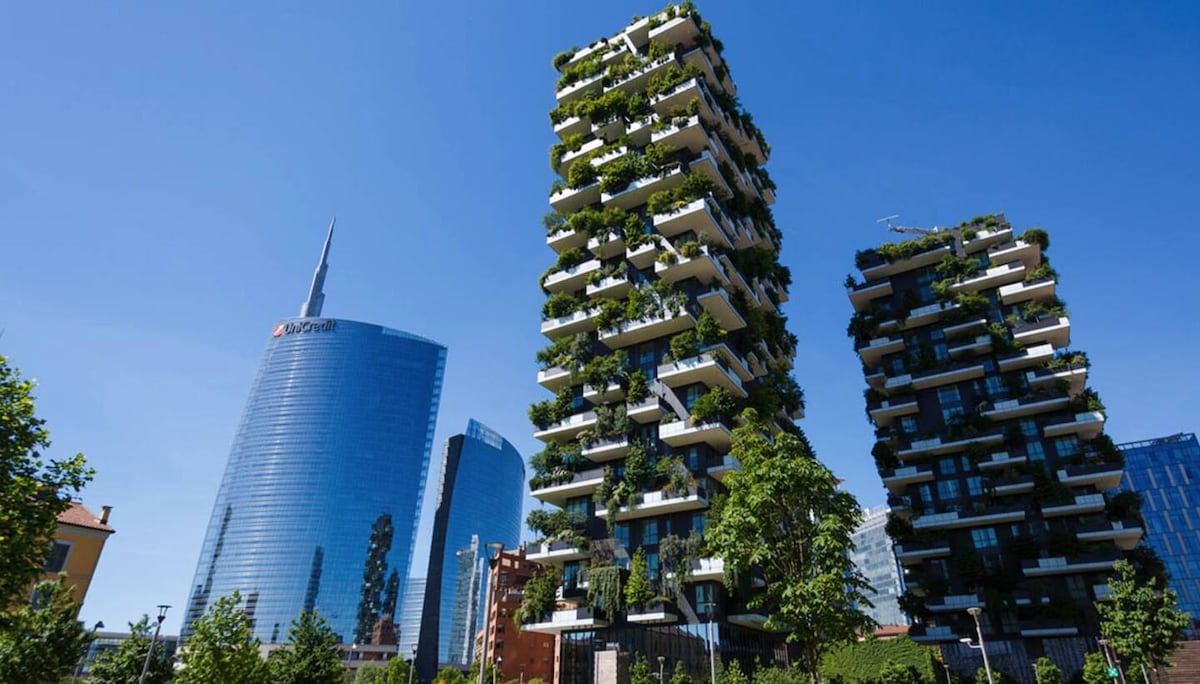 Two modern residential towers are depicted, featuring green terraces filled with vegetation. The sleek glass facade of a nearby skyscraper is visible in the background under a clear blue sky, emphasizing the urban landscape of the area.