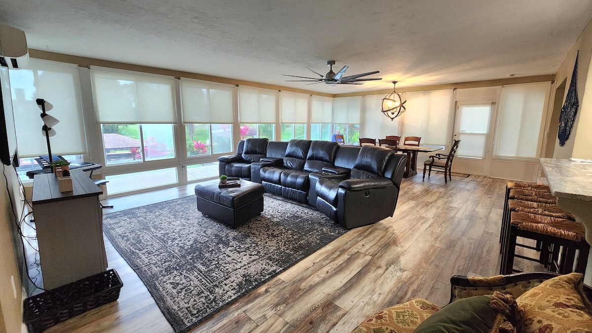 A spacious living area features a black sectional sofa oriented towards a flat-screen television. Large windows adorned with sheer shades allow natural light to fill the space. A dining table with chairs is visible in the background, complemented by decorative elements on the walls.