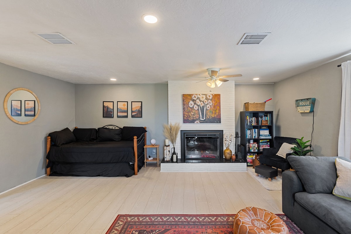 A bright living room features a daybed with black bedding and colorful wall art above. A fireplace with a decorative surround serves as a focal point, while a bookshelf filled with books stands to the side. A cozy chair and decorative elements add warmth to the space.