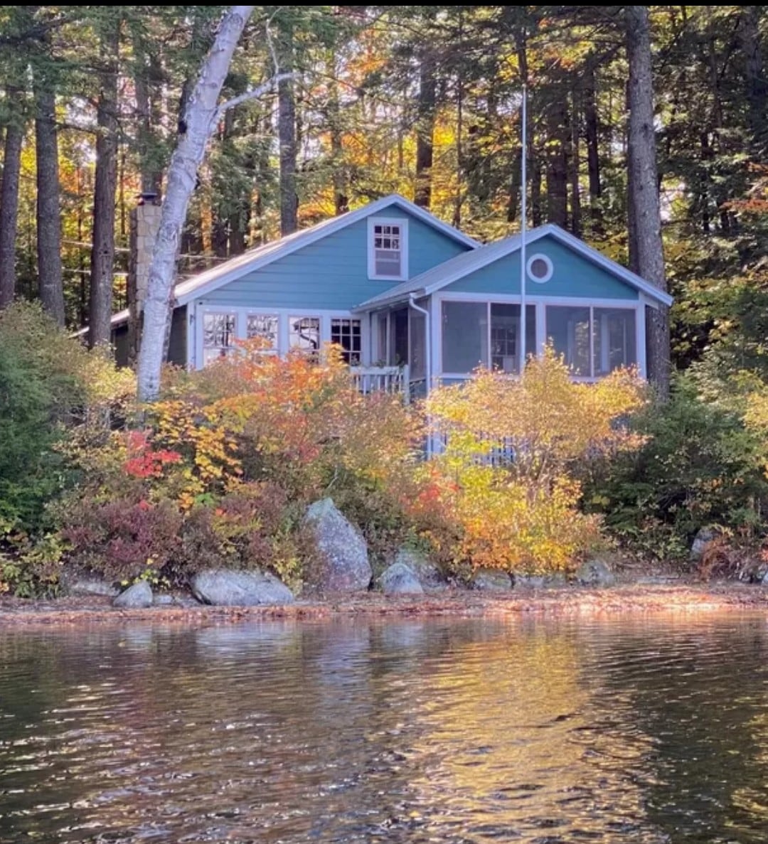 A cozy cabin sits nestled among colorful autumn foliage, reflecting on the calm lake surface. The exterior features a charming blue facade and a screened porch, surrounded by trees and boulders leading to the water’s edge.