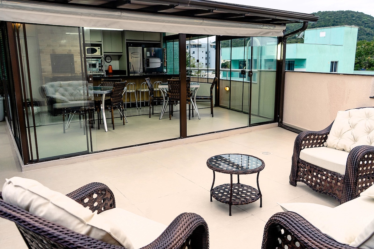 A spacious outdoor terrace is presented, featuring comfortable seating arrangements with wicker furniture. A glass table is placed in the center, while a covered area with a dining table and chairs can be seen through expansive glass walls that allow natural light.