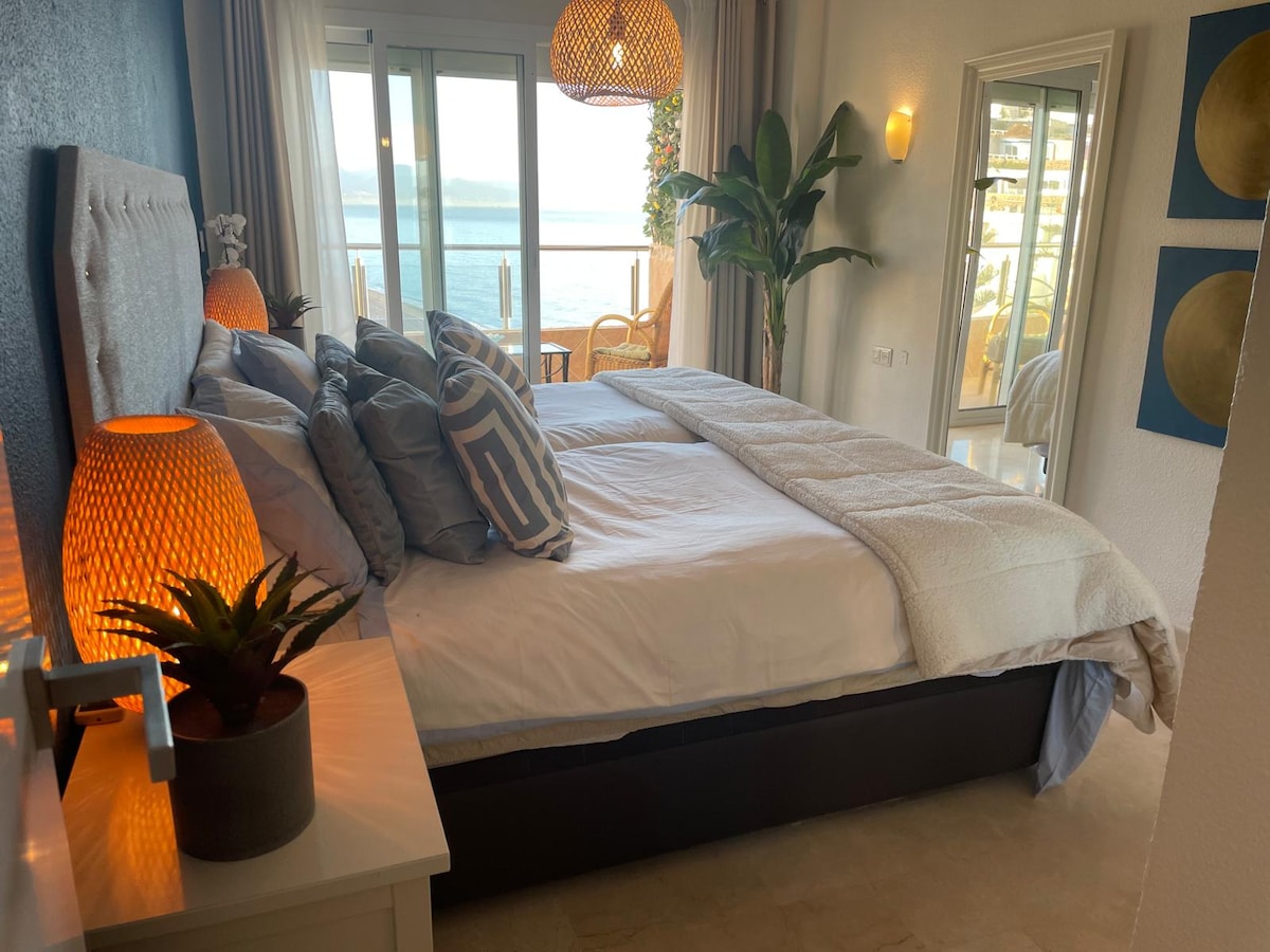 A cozy bedroom features a spacious bed with plush pillows and a soft throw. Warm lamps provide gentle lighting, while a large mirror and a decorative plant add elegance. Sliding glass doors offer a view of the outdoor seating area and beautiful sea landscape.